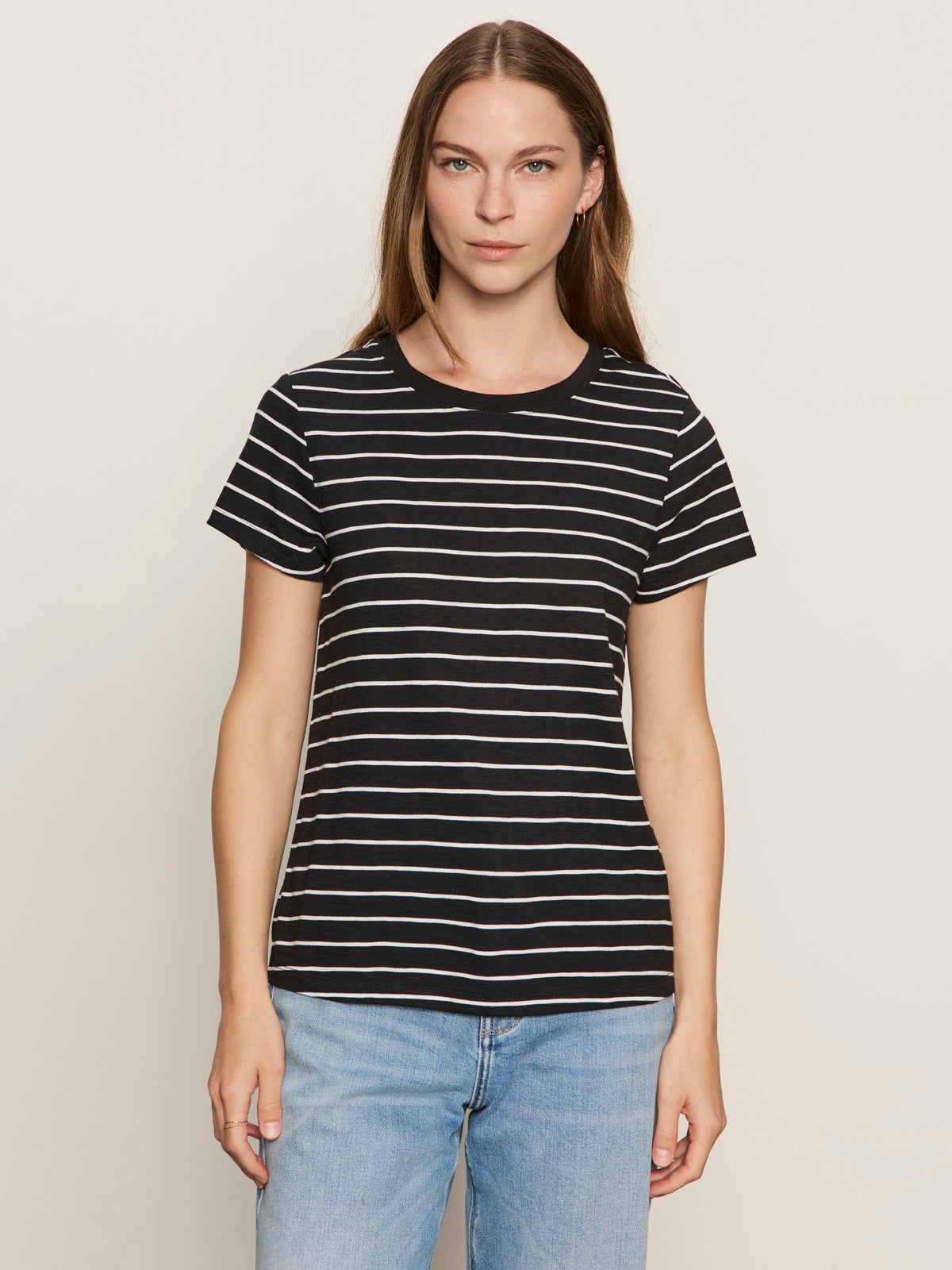 A woman with long brown hair faces forward against a plain background, wearing Sanctuary Clothing’s "the perfect tee white black stripe 5" and light blue jeans. She has a neutral expression.
