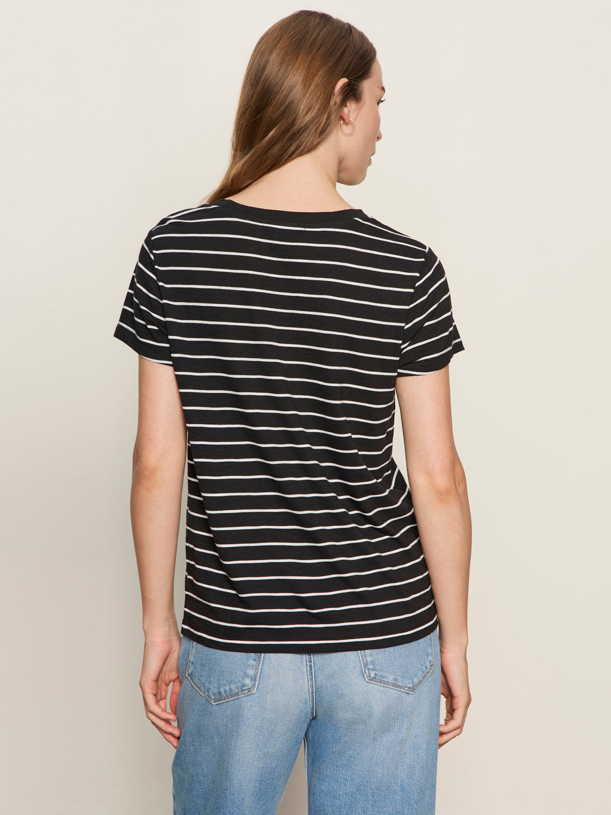 A person with long brown hair stands against an off-white background, wearing Sanctuary Clothing's the perfect tee white black stripe 5 and light blue jeans.