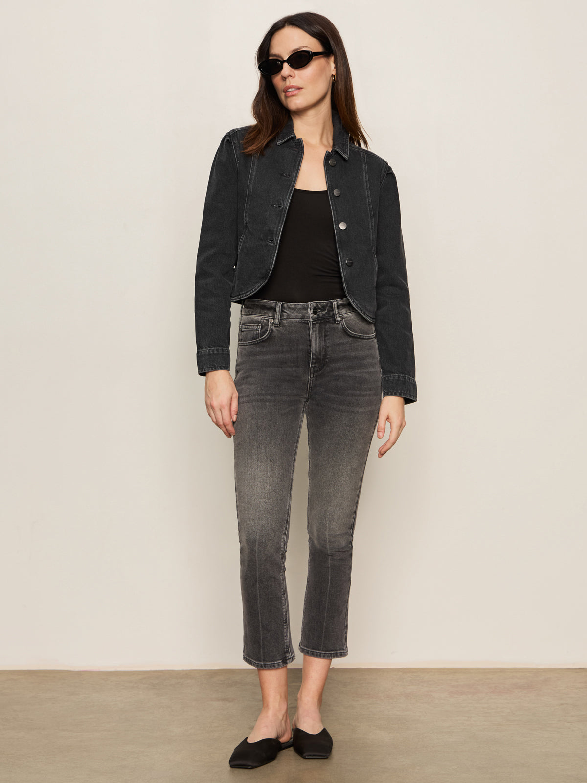 A woman wearing black sunglasses and a Sanctuary Clothing denim jacket in washed black denim, paired with a black top, gray high-waisted jeans, and black flats, stands against a plain light-colored wall.