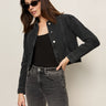 A woman wearing Sanctuary Clothing’s washed black denim jacket stands against a light background, pairing it with black sunglasses, a black top, and high-waisted black jeans; one hand rests on her hip as she tilts her head slightly.