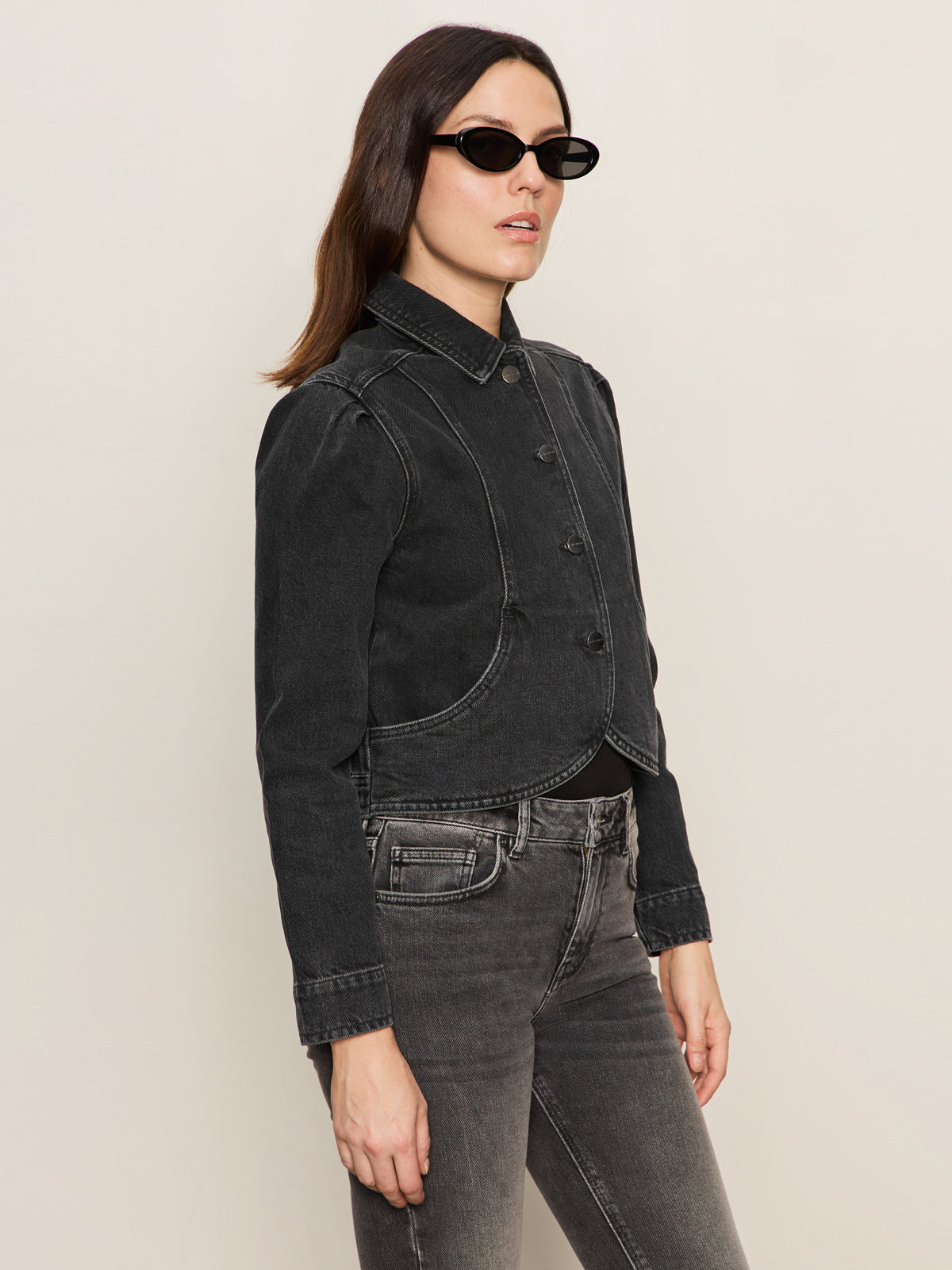 A woman in Sanctuary Clothing's denim jacket washed black denim, paired with black oval sunglasses and matching black jeans, stands against a plain beige background.