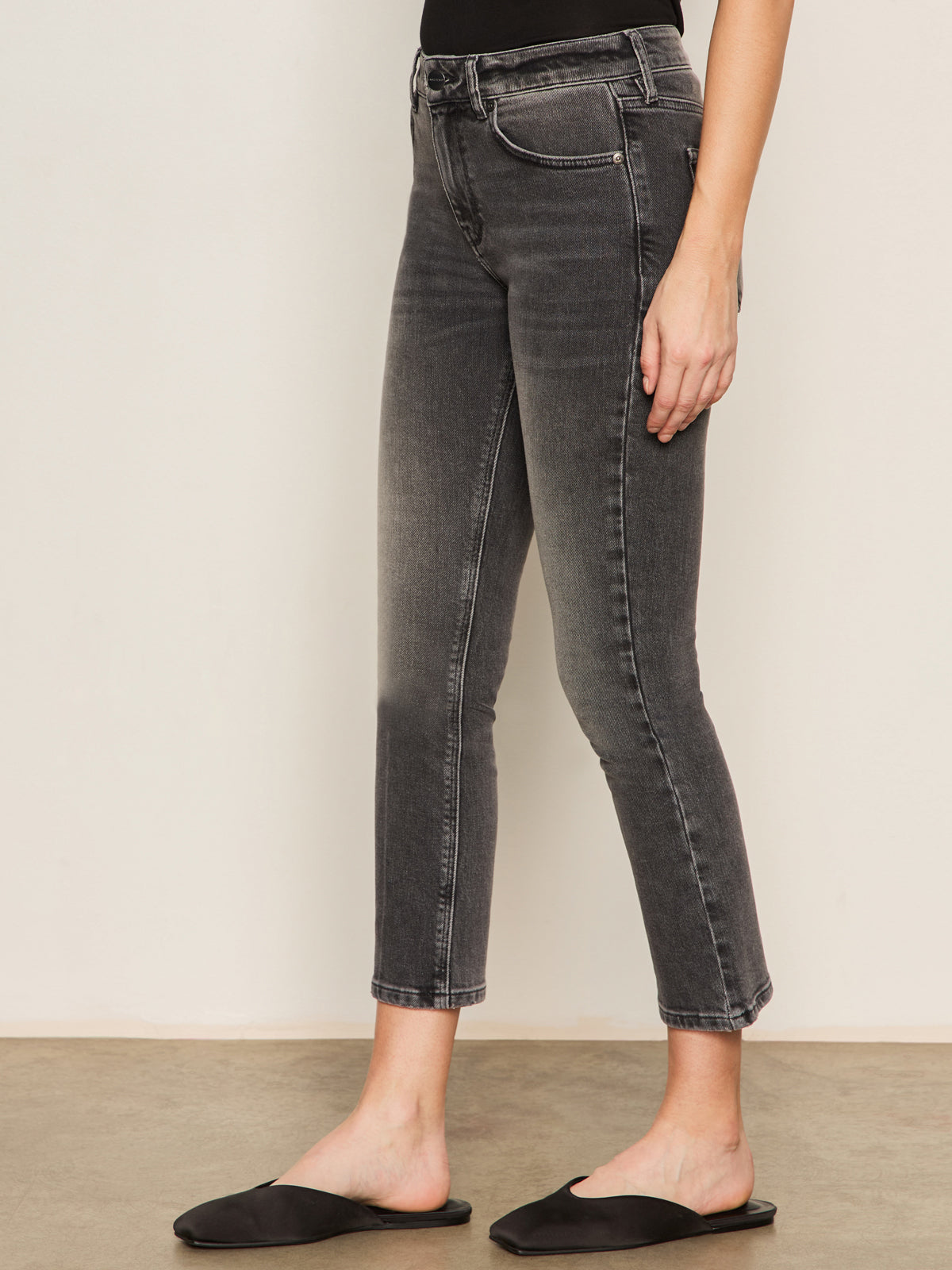 Someone wearing Sanctuary Clothing's astra kick crop jean in washed black denim and black slip-on shoes stands sideways against a plain wall.