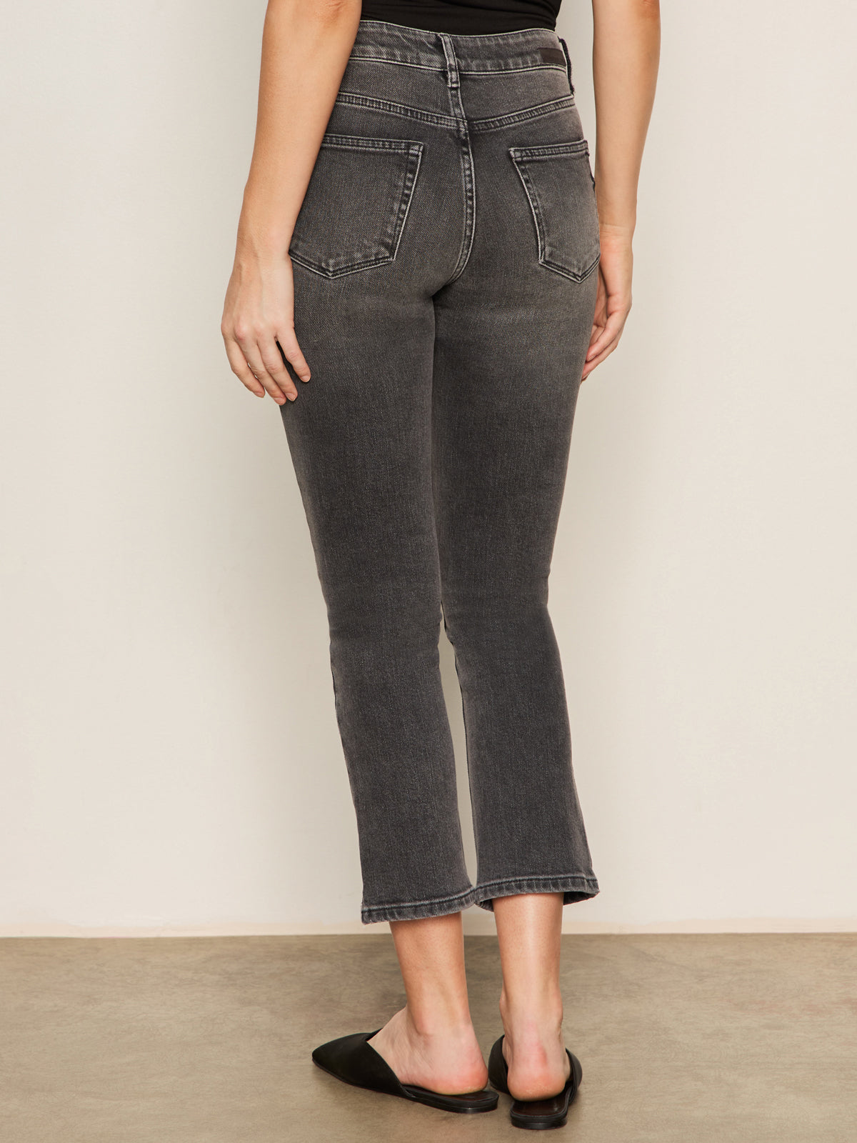 A person faces away, wearing Sanctuary Clothing's astra kick crop jean in washed black denim—high-waisted with raw hems and back pockets—paired with a black sleeveless top and slip-on shoes against a light, plain background.