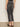 A person faces away, wearing Sanctuary Clothing's astra kick crop jean in washed black denim—high-waisted with raw hems and back pockets—paired with a black sleeveless top and slip-on shoes against a light, plain background.