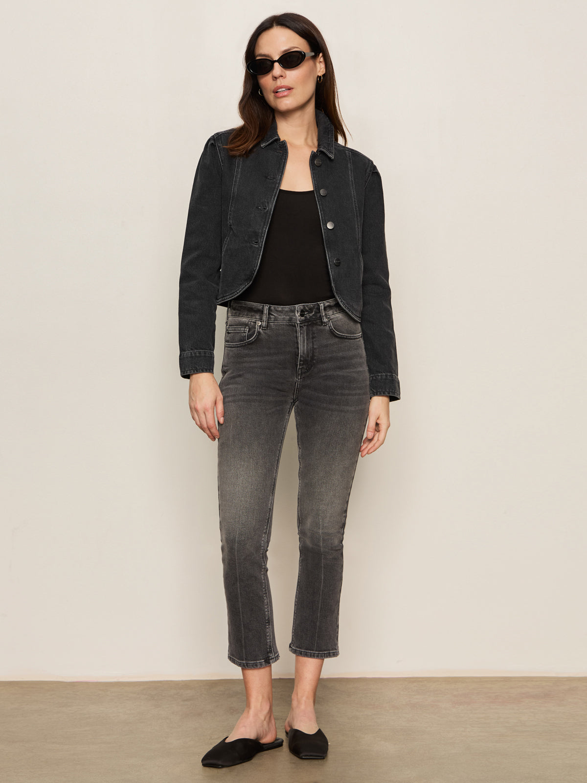 A woman with loose hair in a relaxed pose wears black sunglasses, a cropped black denim jacket, a black top, and Sanctuary Clothing's astra kick crop jean in washed black denim, paired with black slip-on shoes against a plain background.