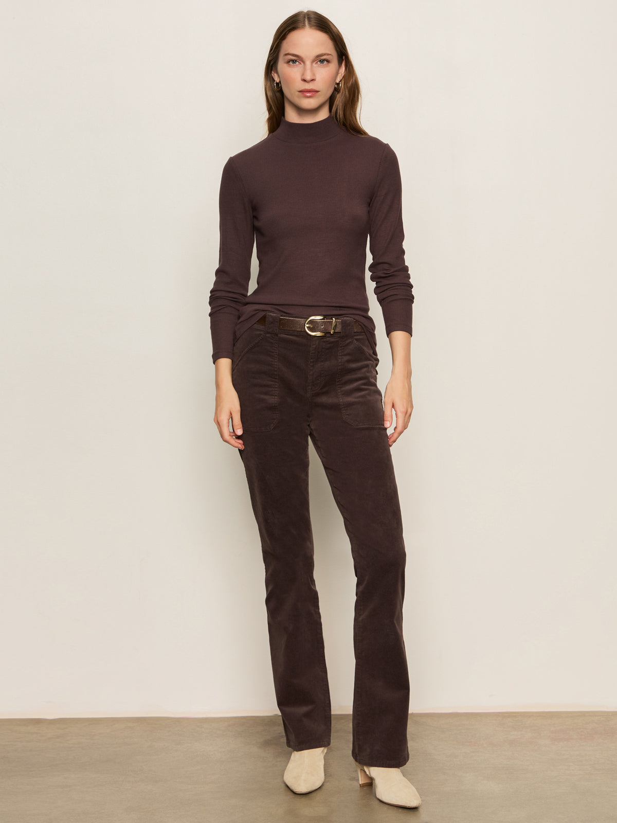 A woman stands against a plain beige wall, wearing the Sanctuary Clothing ls mock essential top semi sweet, matching dark brown belted trousers, and light-colored shoes. She faces forward with a neutral expression.