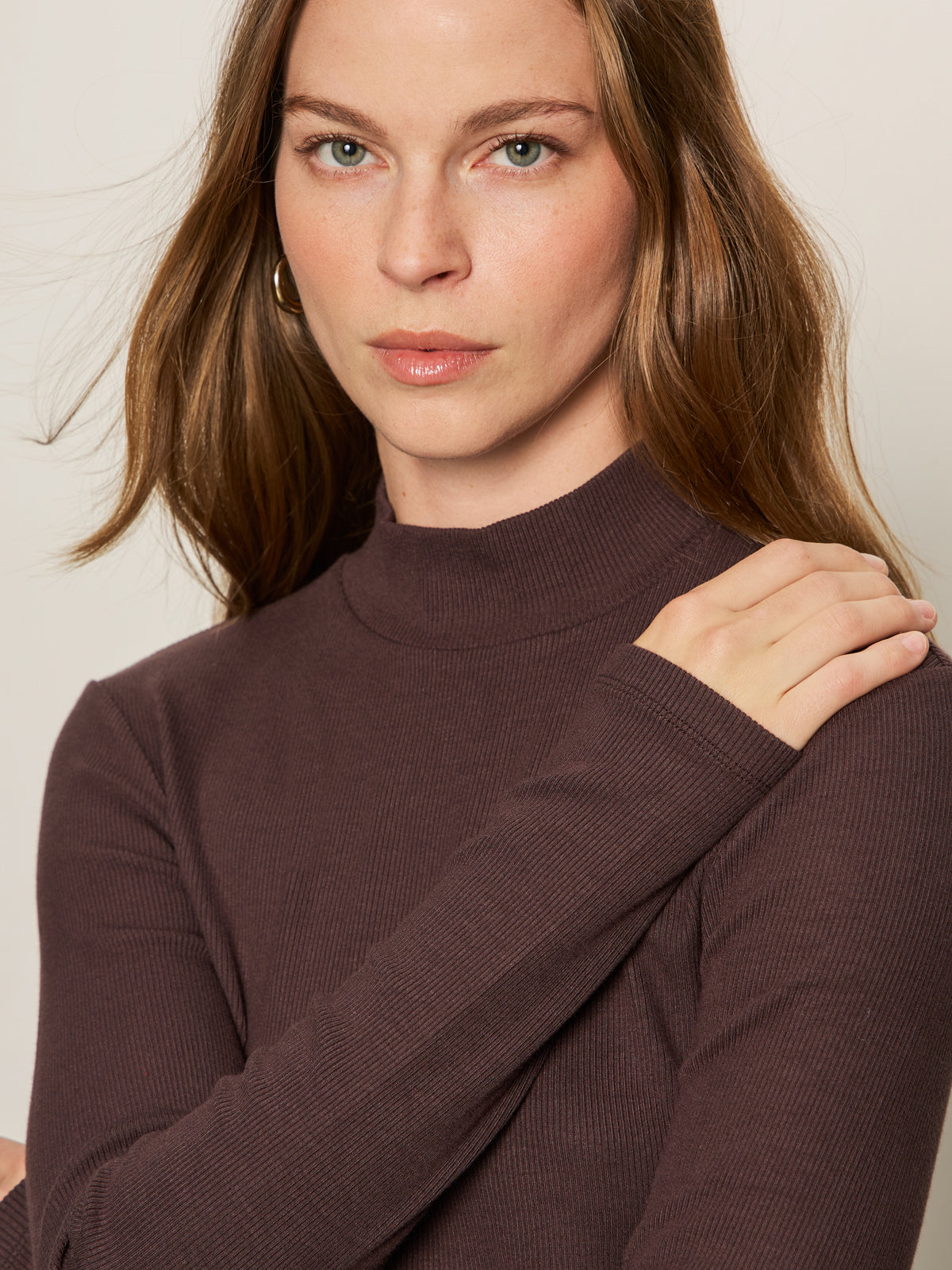 A woman with long brown hair wears the Sanctuary Clothing ls mock essential top in semi sweet, looking at the camera with her left hand on her right shoulder against a neutral background.