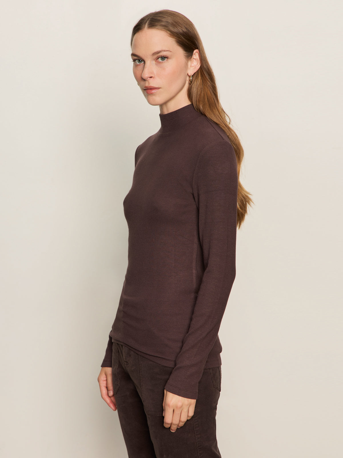 A woman with long brown hair models the Sanctuary Clothing ls mock essential top in semi sweet, standing sideways against an off-white background and looking towards the camera with a neutral expression.