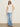A woman wears Sanctuary Clothing's side tie muscle tee in white with light blue wide-leg jeans, black sandals, and sunglasses, standing against a plain beige background.