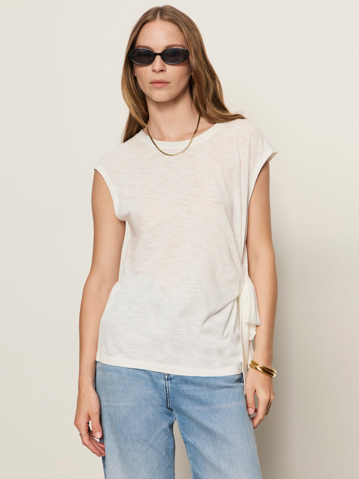 A woman stands against a plain light background wearing Sanctuary Clothing's Side Tie Muscle Tee in white, styled with sunglasses, a gold chain necklace, bracelets, and light blue jeans.