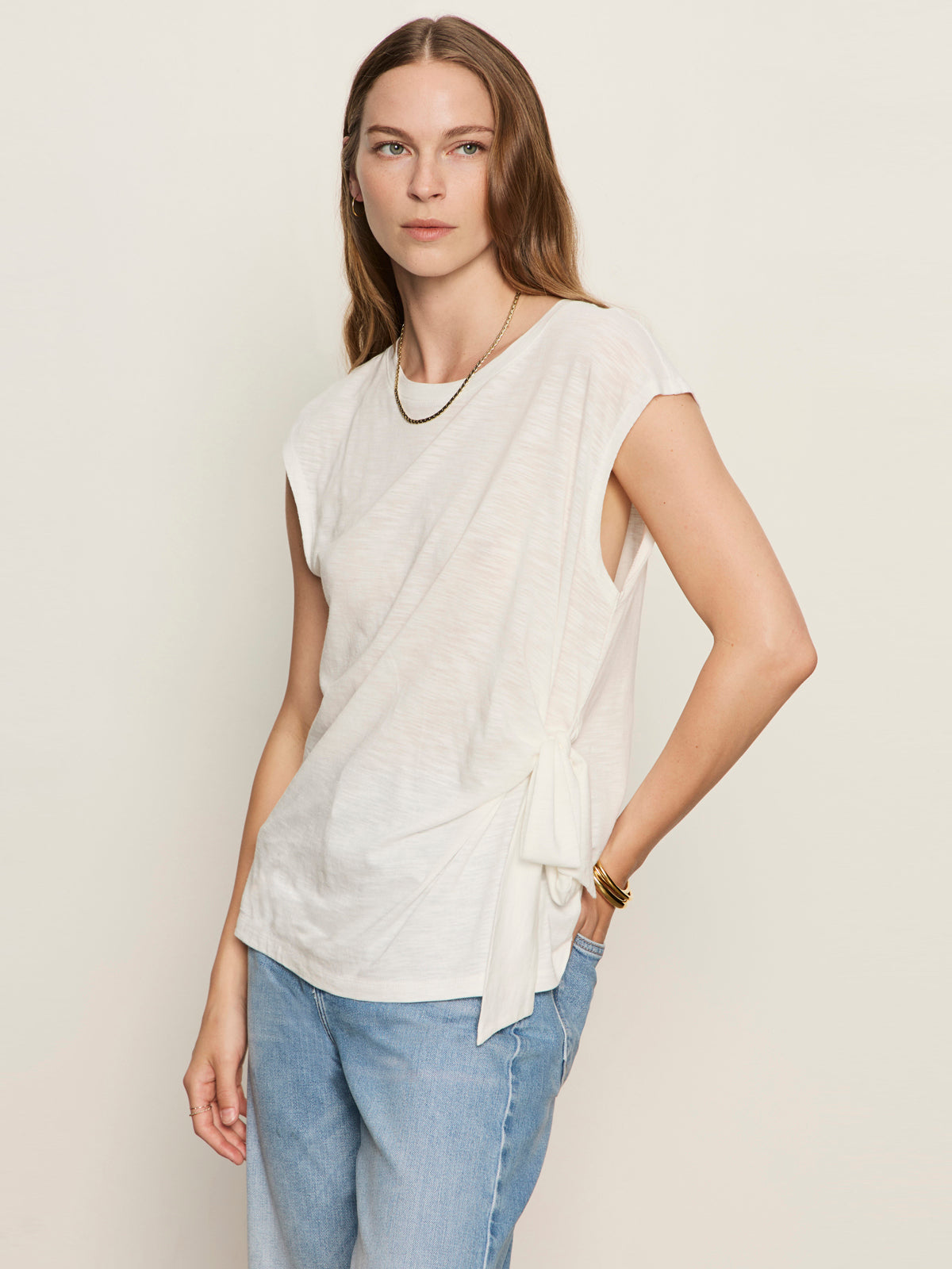 A woman with long brown hair models the Sanctuary Clothing side tie muscle tee in white, paired with light blue jeans and gold jewelry, set against a plain light background.