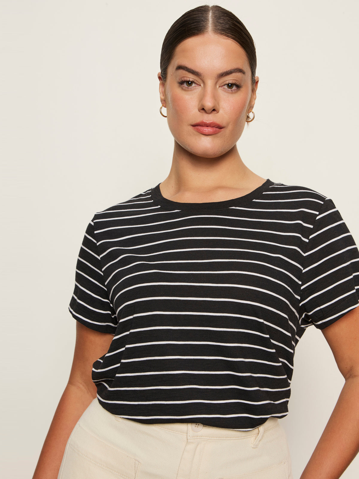 A woman with dark hair pulled back wears gold hoop earrings and the Sanctuary Clothing “the perfect tee white black stripe 6,” standing against a plain light background.
