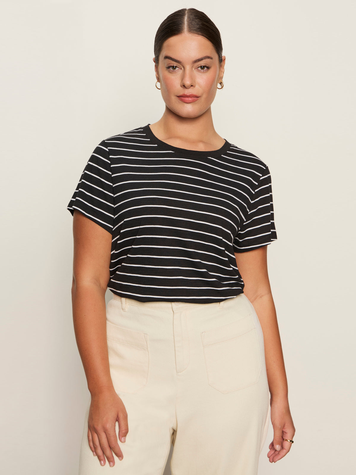 A woman with dark hair pulled back and small hoop earrings wears the Sanctuary Clothing "the perfect tee white black stripe 6," a black and white striped t-shirt, tucked into high-waisted, light-colored pants against a plain background.