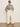 A woman wearing Sanctuary Clothing's "the perfect tee green oasis" (extended sizing), cream wide-leg pants, and sandals stands against a plain off-white wall with her hands in her pockets.