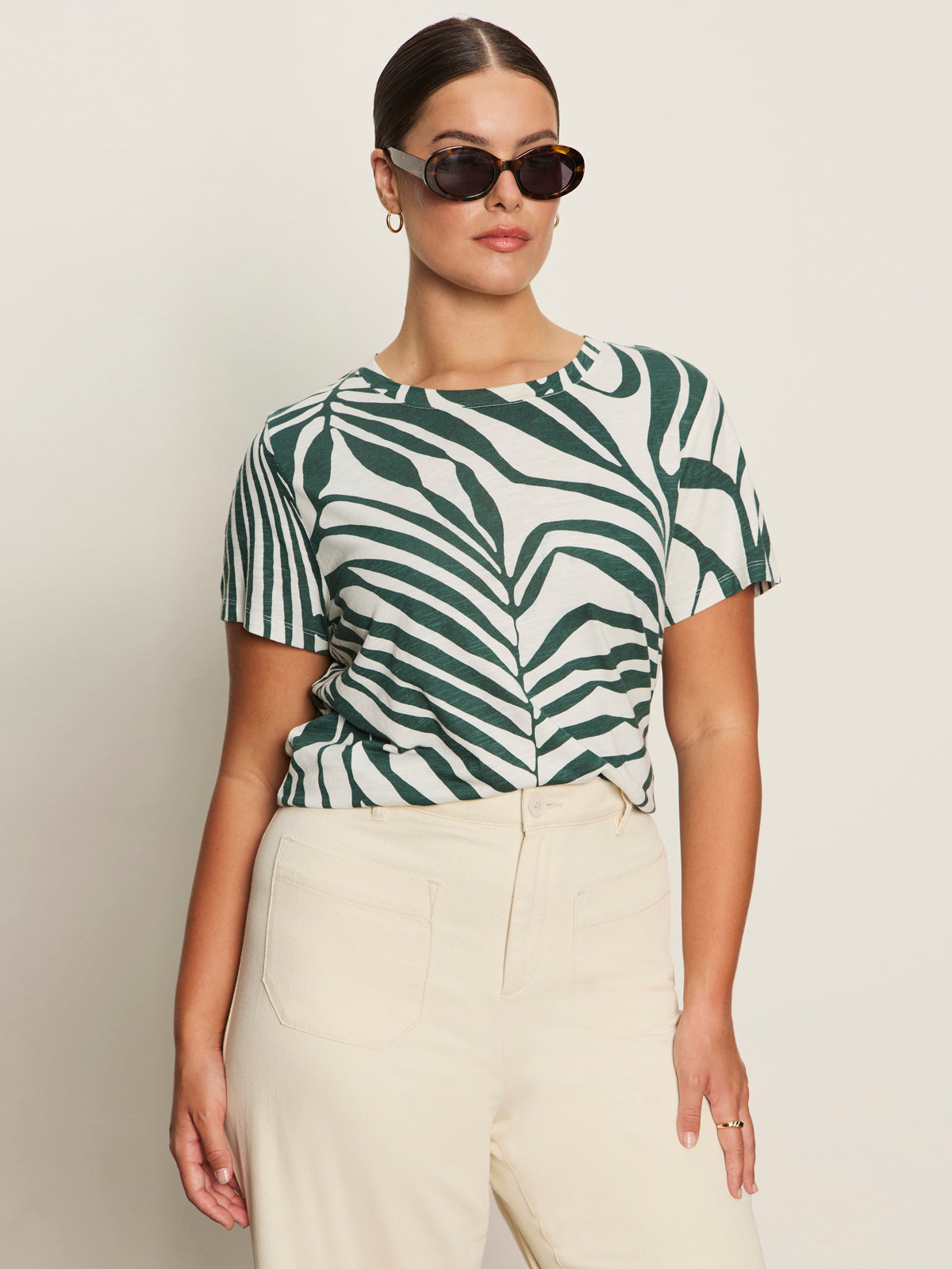 A woman wearing Sanctuary Clothing's the perfect tee green oasis (extended sizing) and high-waisted cream pants stands against a light background, with one hand in her pocket.