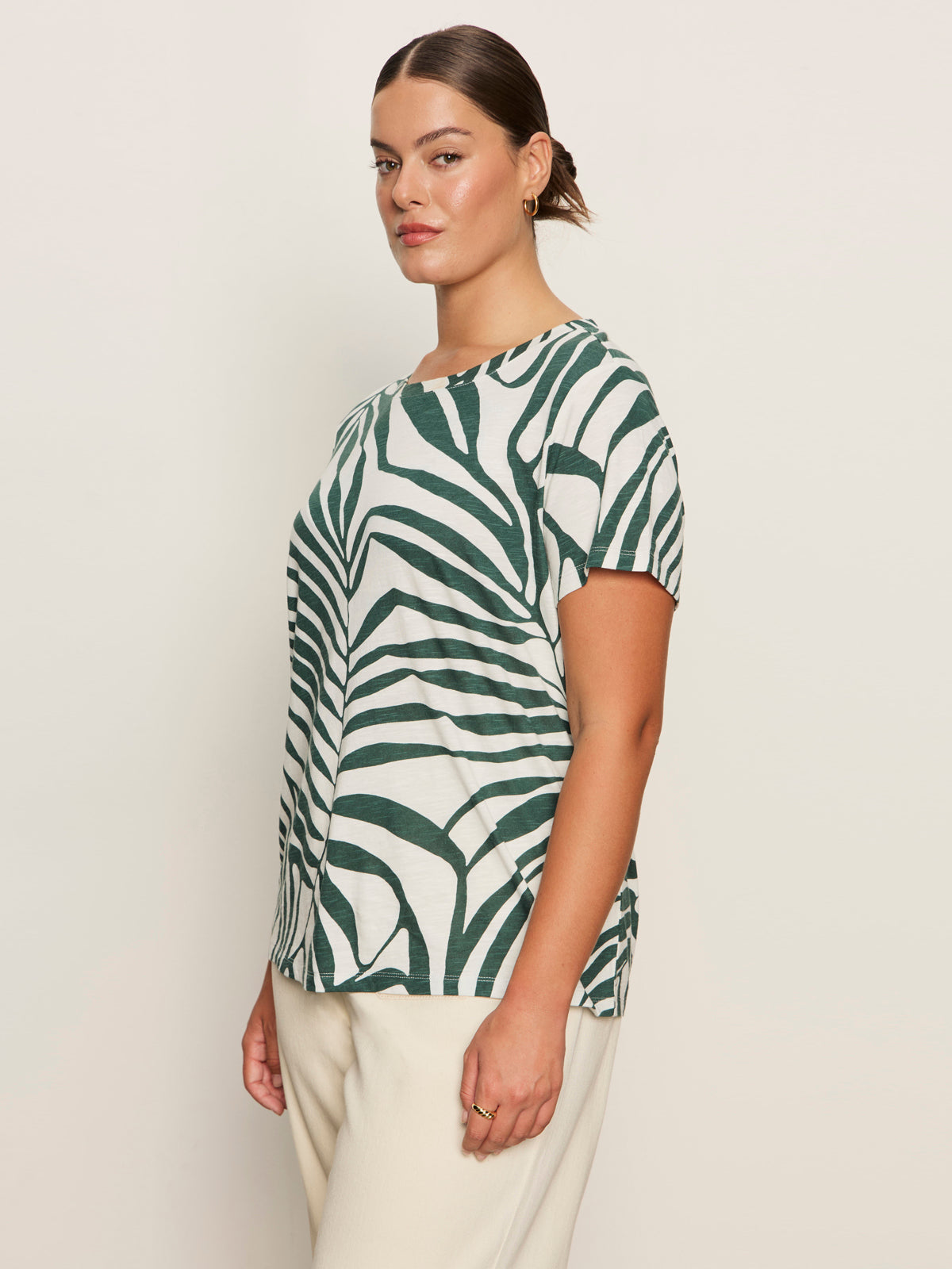 A woman stands sideways against a plain light background wearing Sanctuary Clothing’s “the perfect tee green oasis extended sizing,” featuring bold green leaf patterns, paired with cream pants and gold hoop earrings.