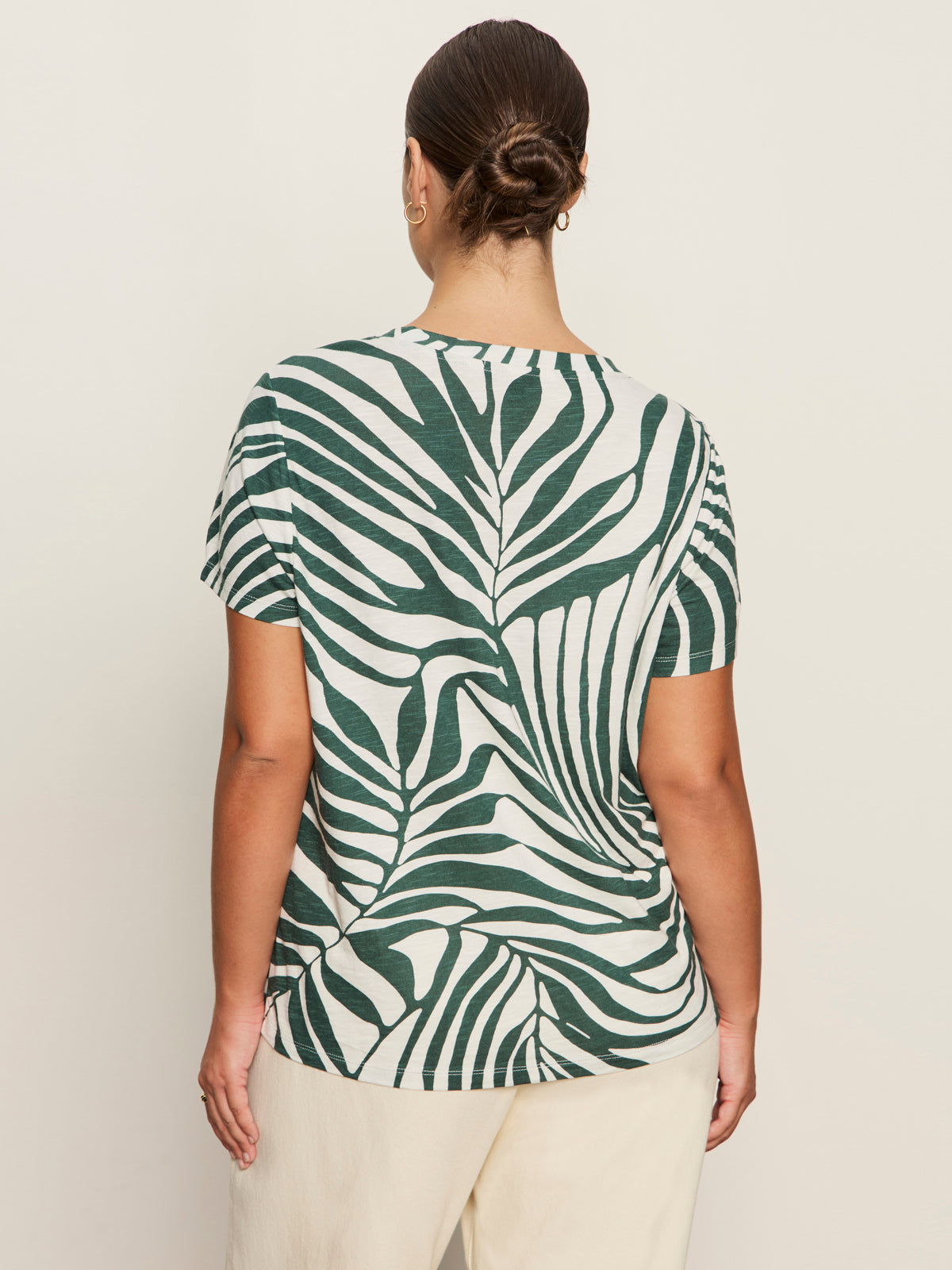 A person with dark hair in a low bun wears Sanctuary Clothing's the perfect tee green oasis extended sizing with cream pants, shown from the back against a plain light background.