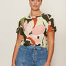 A woman with dark hair tied back wears Sanctuary Clothing’s the perfect tee reef vibes extended sizing, tucked into high-waisted blue jeans. She stands against a light background, looking confidently at the camera.