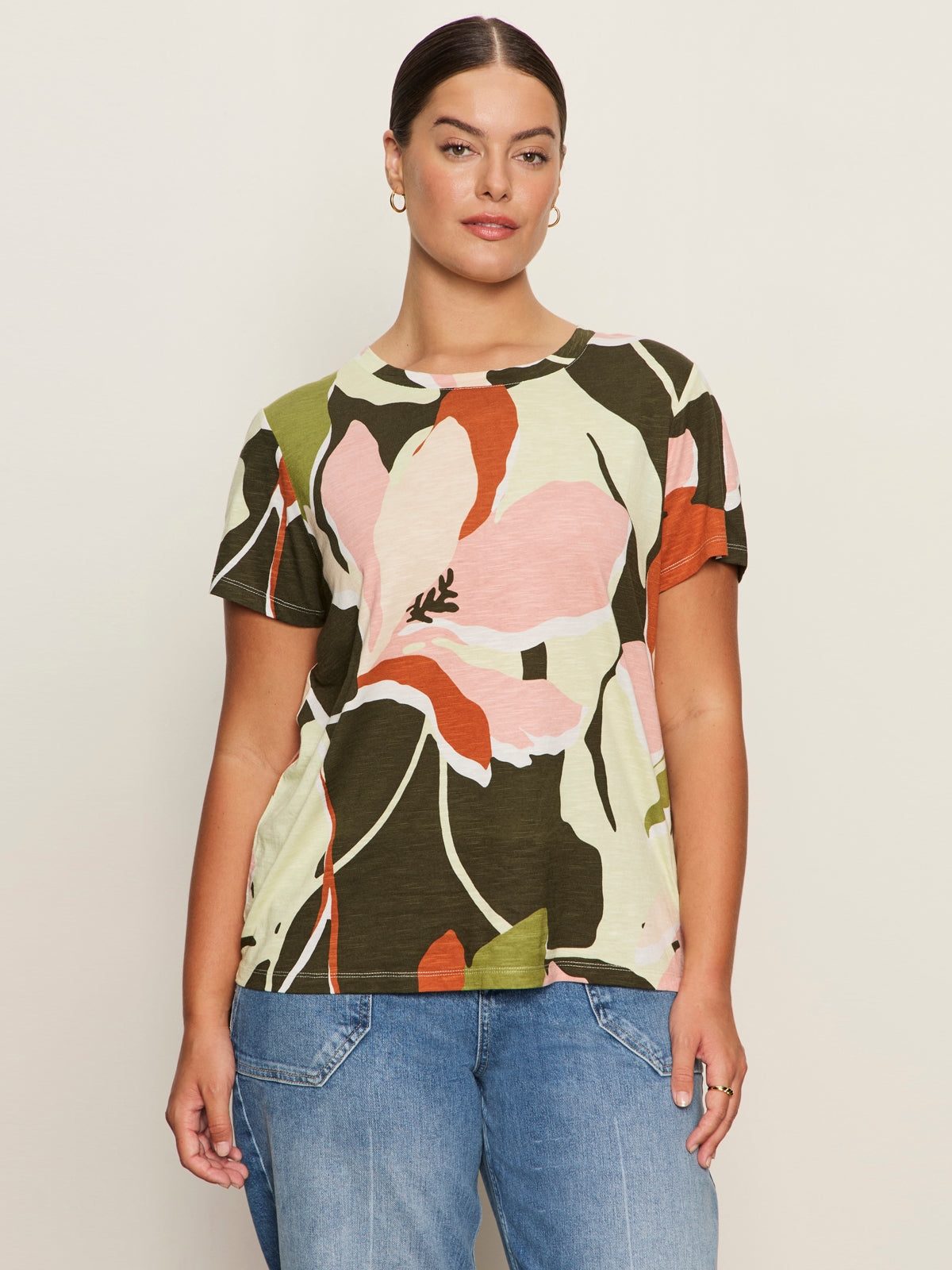 A woman with dark hair tied back wears Sanctuary Clothing's "the perfect tee reef vibes extended sizing" in a floral pink, green, and orange print with gold hoop earrings and blue jeans, standing against a plain off-white background.