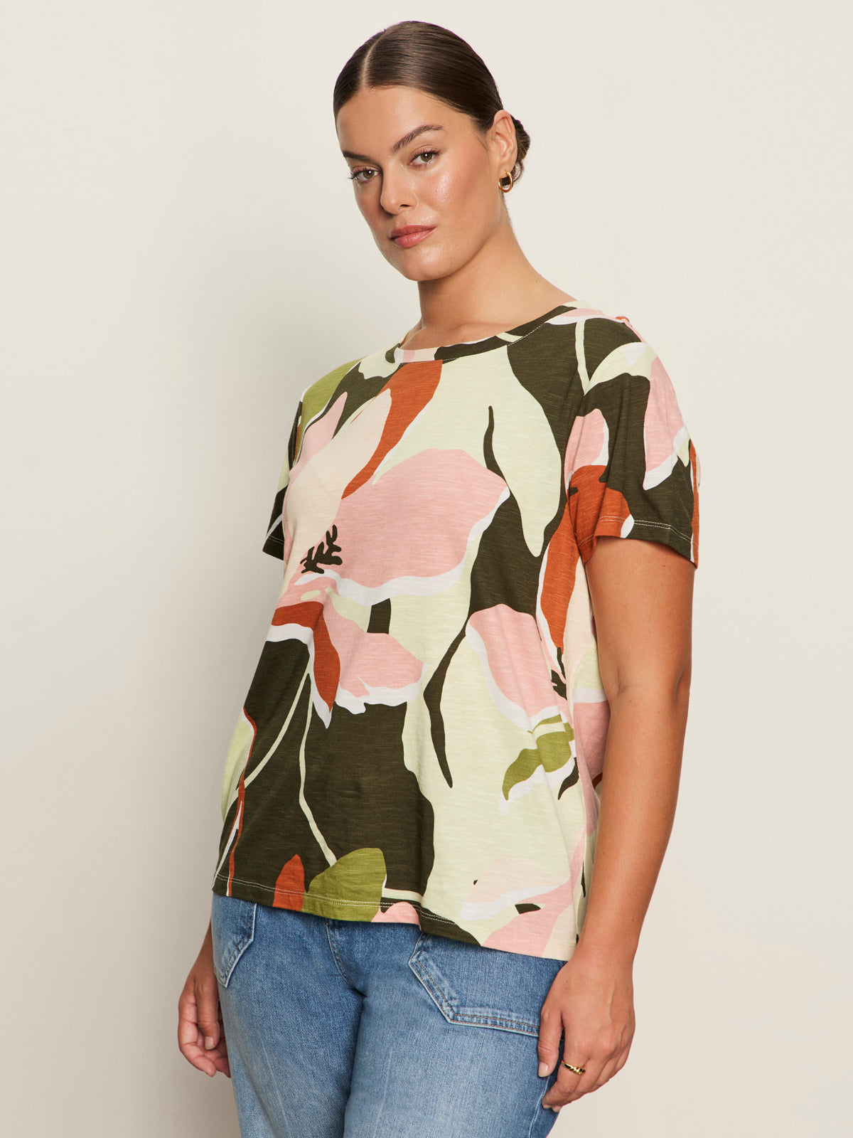 A woman with brown hair in a slicked-back style wears the Sanctuary Clothing "the perfect tee reef vibes extended sizing," featuring pink, orange, green, and black florals, along with blue jeans, standing before an off-white background.