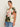 A woman with brown hair in a slicked-back style wears the Sanctuary Clothing "the perfect tee reef vibes extended sizing," featuring pink, orange, green, and black florals, along with blue jeans, standing before an off-white background.