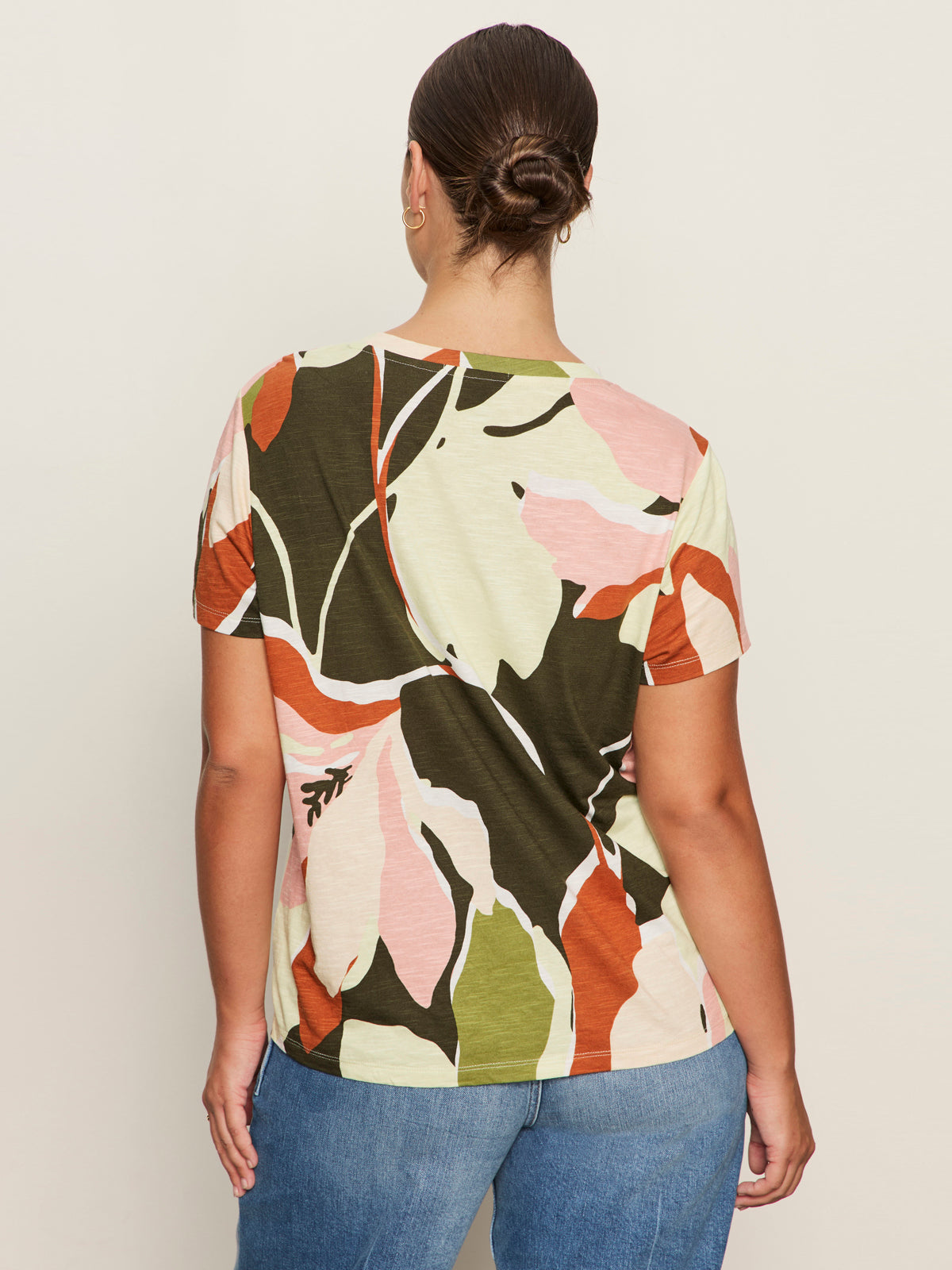 A woman with dark hair in a bun wears the Sanctuary Clothing "the perfect tee reef vibes extended sizing," a short-sleeve top with a colorful abstract floral pattern, and blue jeans, standing facing away from the camera against a plain background.