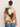 A woman with dark hair in a bun wears the Sanctuary Clothing "the perfect tee reef vibes extended sizing," a short-sleeve top with a colorful abstract floral pattern, and blue jeans, standing facing away from the camera against a plain background.