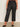 Someone wearing the Marine Jeans After Hours (extended sizing) by Sanctuary Clothing, featuring black cropped wide-leg style with front patch pockets, and black pointed-toe flats, stands against a light wall and concrete floor.