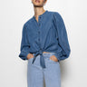 A person poses against a white background wearing the Sanctuary Clothing tie front volume blouse in blue slate and light blue jeans, with their hands in their pockets, looking forward.