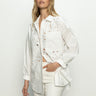 A woman with long blonde hair is wearing Sanctuary Clothing's lace boyfriend tie back shirt in white, paired with white pants. She stands against a light background, confidently looking at the camera.