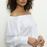 A woman with curly hair is wearing a Sanctuary Clothing ballet tie top in white, paired with light-colored pants, standing against a plain background.