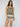 A woman with long brown hair models the Sanctuary Clothing button back cami in mezzo sage, paired with matching shorts featuring a pink and cream ornate pattern, standing against a plain light gray background.