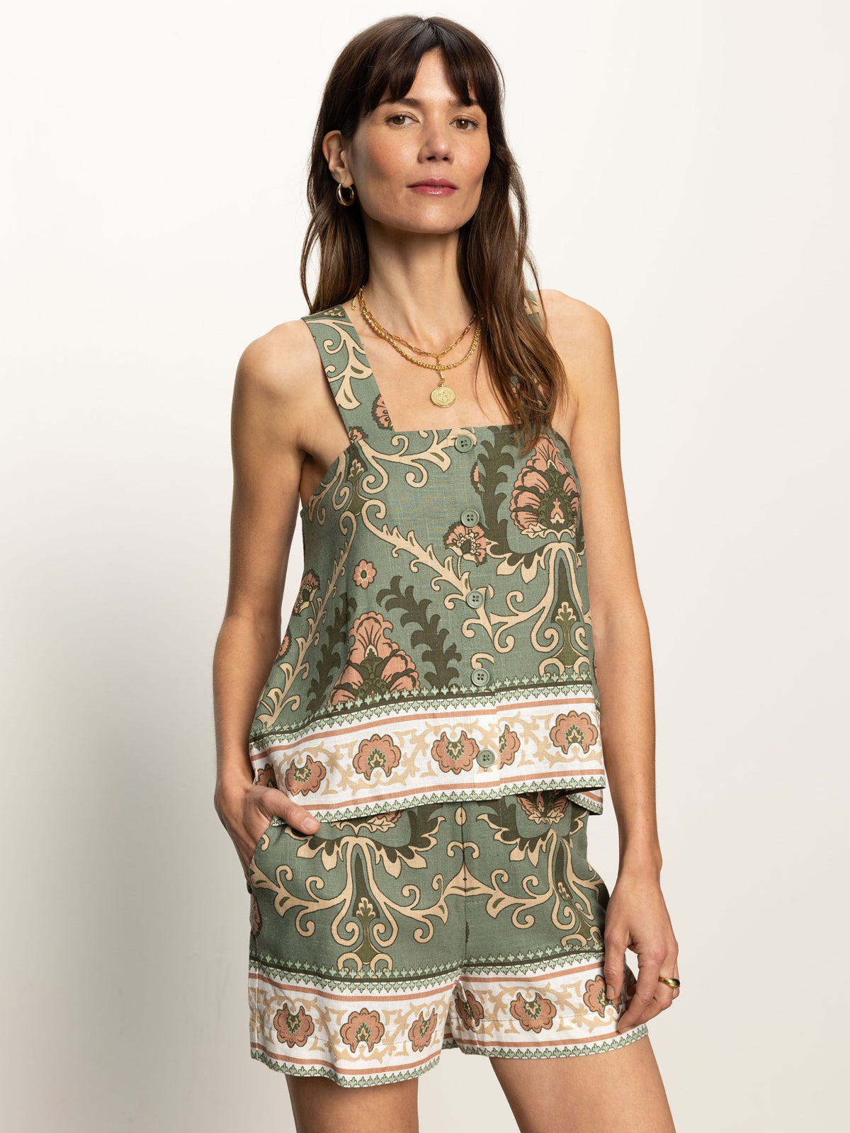 A woman with long brown hair and layered necklaces is wearing the "button back cami mezzo sage" by Sanctuary Clothing, a green patterned sleeveless top with orange and white floral accents, paired with matching shorts. Her left hand rests in her pocket against a neutral background.