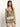 A woman with long brown hair and layered necklaces is wearing the "button back cami mezzo sage" by Sanctuary Clothing, a green patterned sleeveless top with orange and white floral accents, paired with matching shorts. Her left hand rests in her pocket against a neutral background.