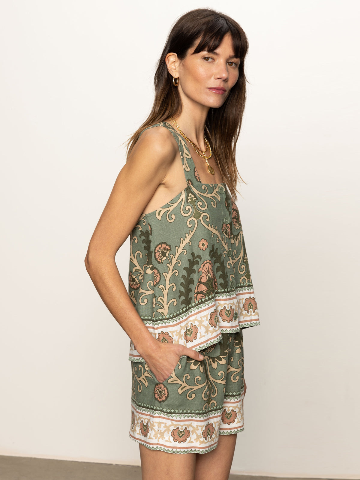 A woman with long brown hair and gold hoop earrings poses sideways, hands in pockets, wearing the Sanctuary Clothing's "button back cami mezzo sage" top and matching shorts against a plain white background.