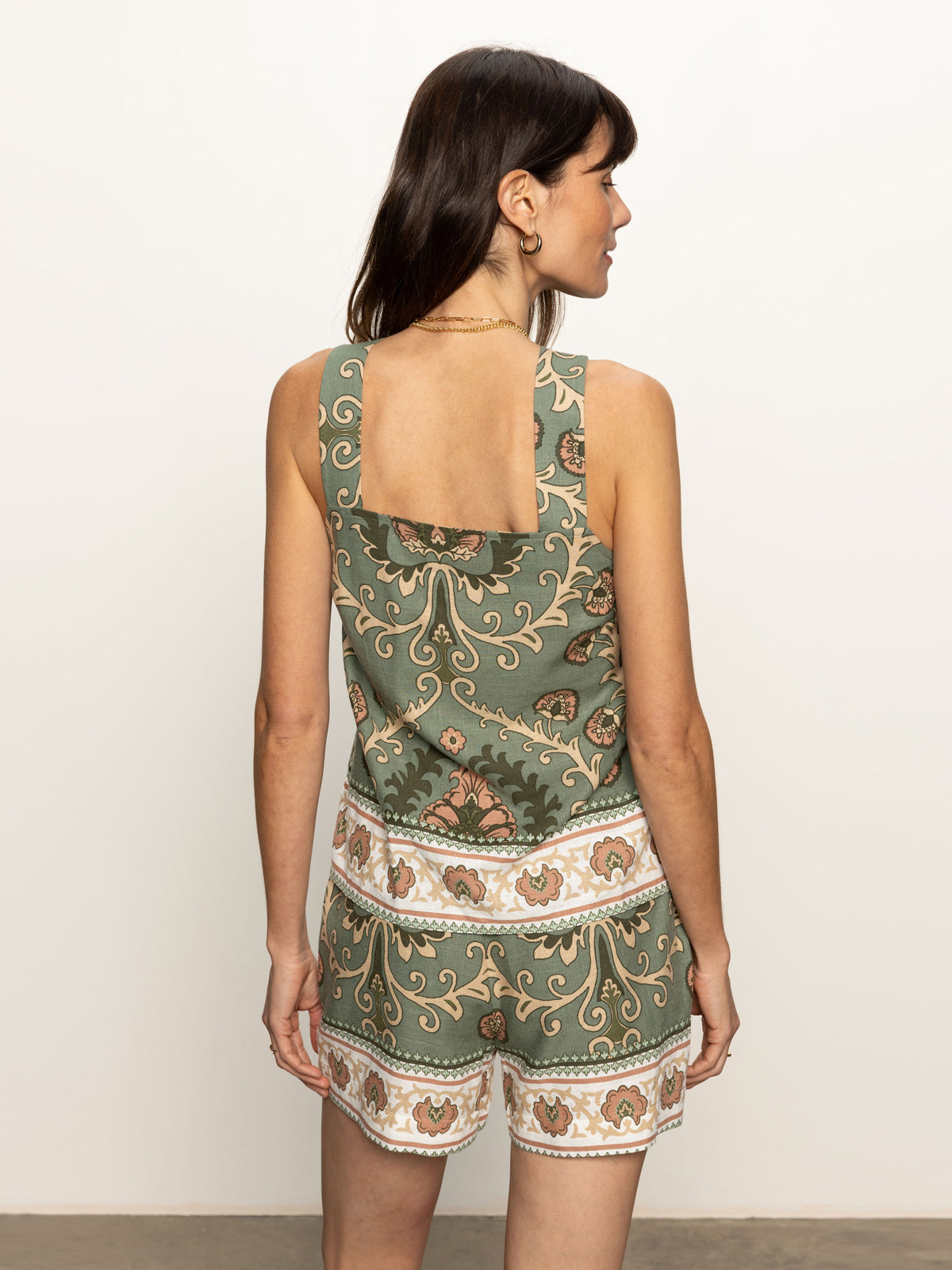 A woman with long dark hair stands against a plain background, wearing the Sanctuary Clothing button back cami in mezzo sage with matching shorts. The ensemble features an intricate floral and geometric pattern in white, pink, and gold as she faces slightly away from the camera.