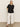 A woman with straight blonde hair stands against a plain light-colored wall, wearing the Sanctuary Clothing relaxed vacation shirt in black, styled with wide-leg white pants and black sandals, hands in her pockets for a laid-back vibe.