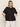 A woman with straight blonde hair wears the Sanctuary Clothing relaxed vacation shirt in black, featuring a loose fit, short sleeves, and a chest pocket, paired with white pants against a plain beige background.