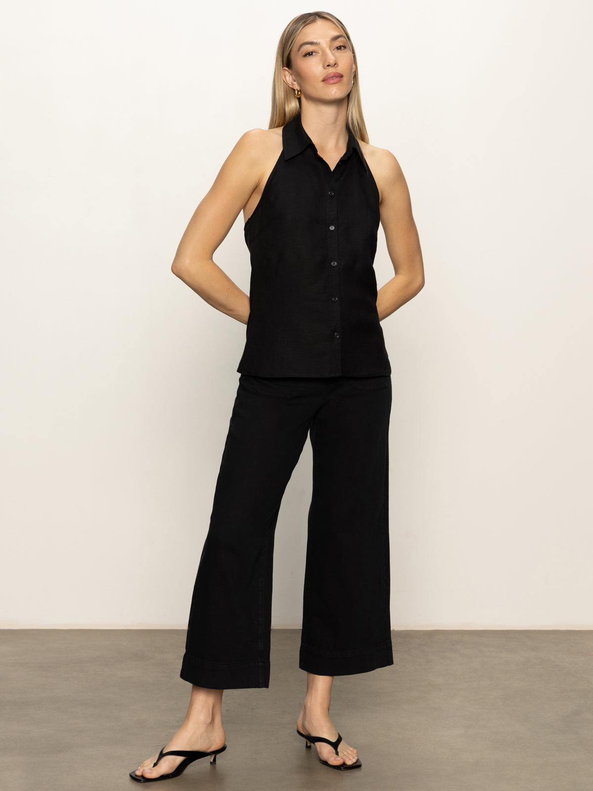 A woman with straight blonde hair poses confidently against a plain background, wearing Sanctuary Clothing’s the long weekend tank in black, paired with black wide-leg pants and black heeled sandals.