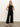 A woman with straight blonde hair poses confidently against a plain background, wearing Sanctuary Clothing’s the long weekend tank in black, paired with black wide-leg pants and black heeled sandals.