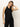 A woman with long blonde hair wears the long weekend tank black by Sanctuary Clothing and black pants, standing against a light background with one hand touching her hair and looking downward.