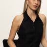 A woman with straight blonde hair and light skin sits on a black chair, wearing Sanctuary Clothing's the long weekend tank in black. The sleeveless, collared button-up top complements her neutral expression as she gazes off to the side.