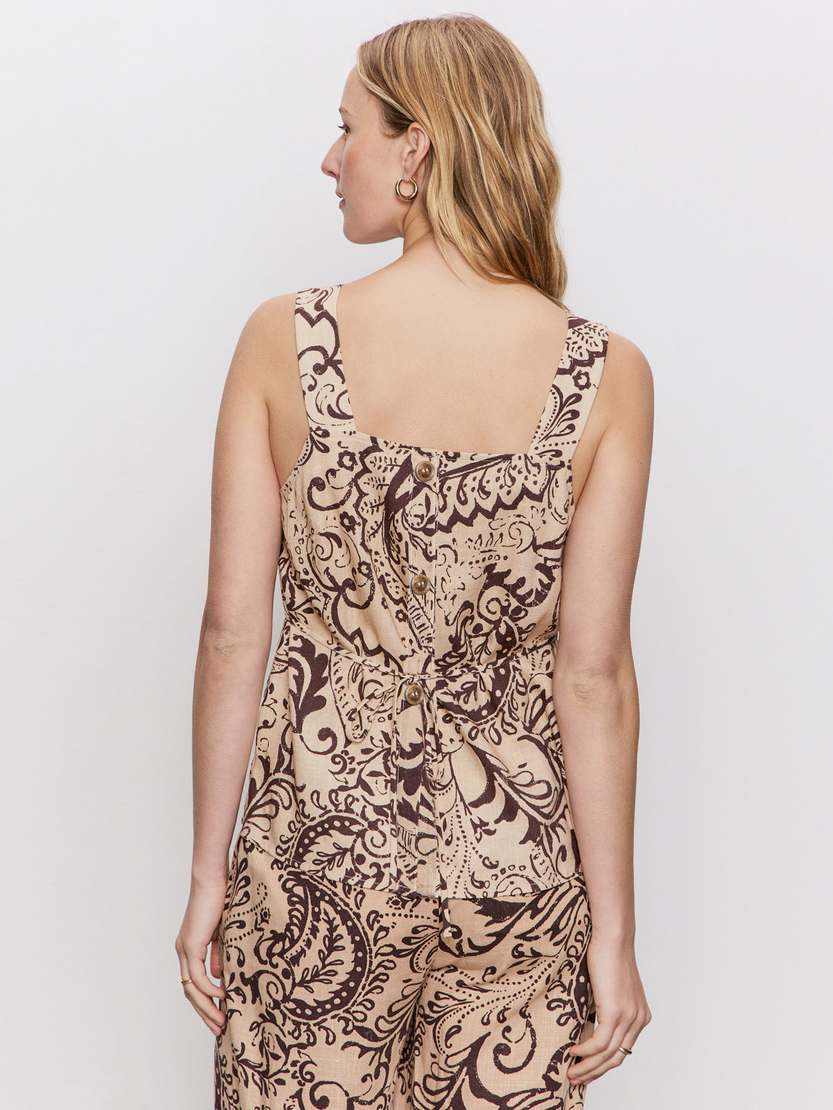A woman with blonde hair models the Sanctuary Clothing desert tank desert bandana, a sleeveless beige jumpsuit with a brown paisley pattern, tie waist, and wide straps. She is shown from the back against a plain white background.