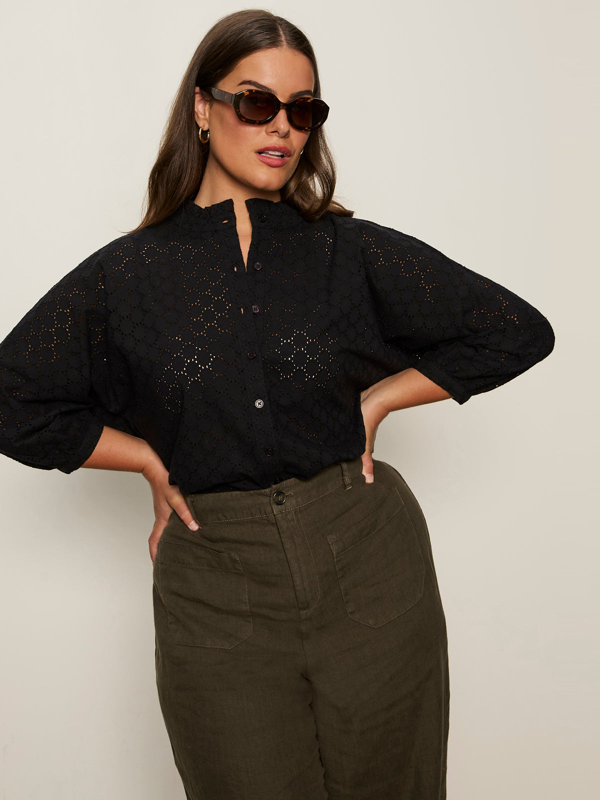 A woman wearing dark sunglasses and the Sanctuary Clothing petal drop blouse black extended sizing stands with her hands on her hips, paired with high-waisted olive green pants against a plain light background.