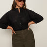 A woman wearing dark sunglasses and the Sanctuary Clothing petal drop blouse black extended sizing stands with her hands on her hips, paired with high-waisted olive green pants against a plain light background.