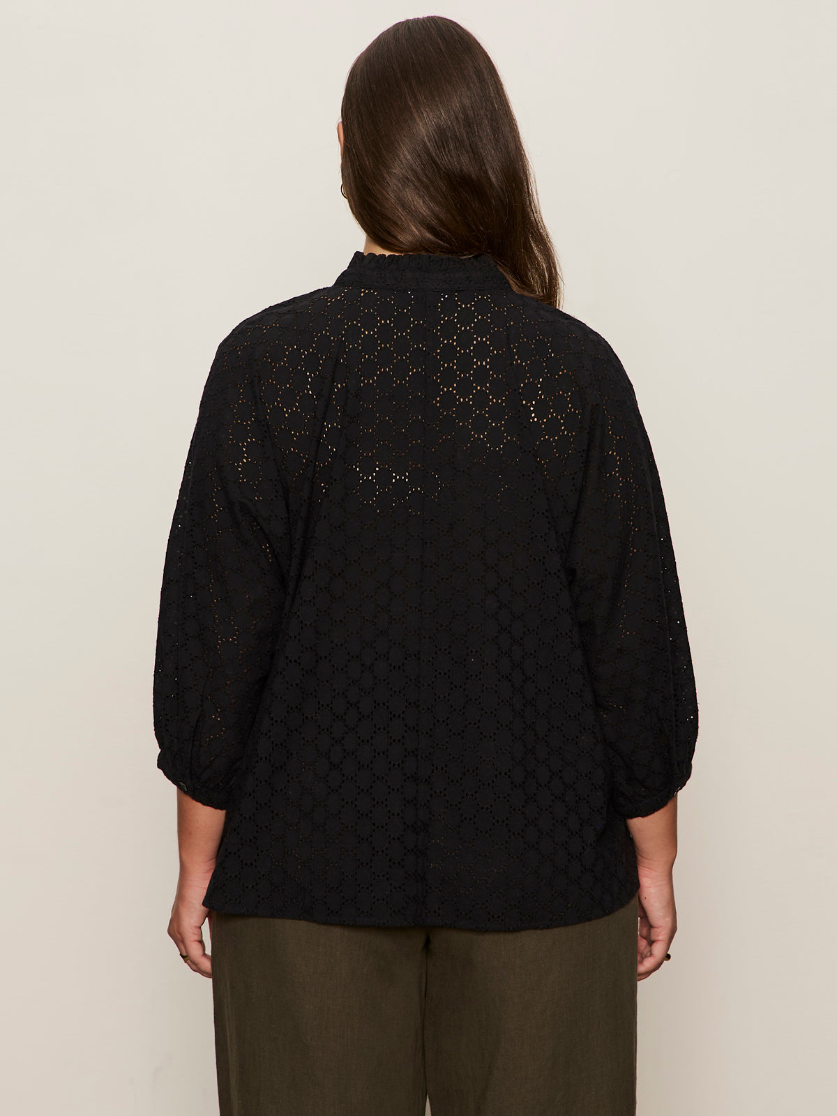 A person with long brown hair, seen from behind, wears the Sanctuary Clothing petal drop blouse black extended sizing and olive green pants against a plain beige background.