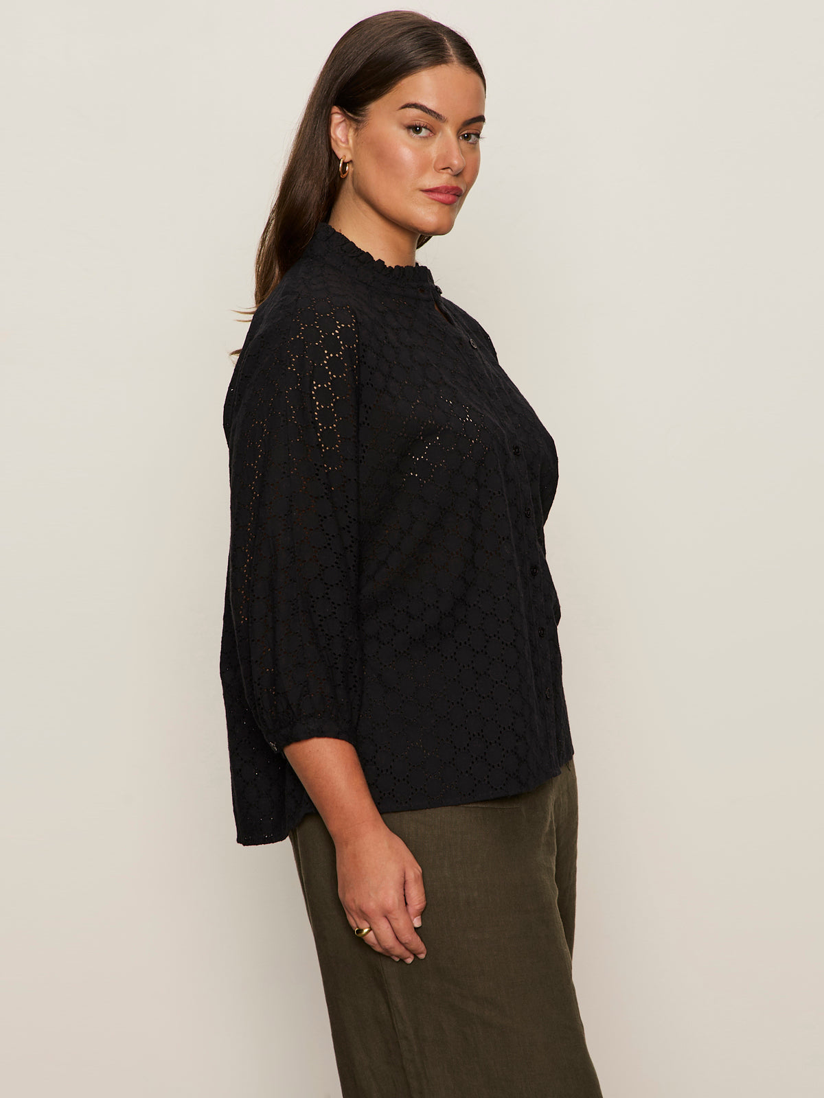 A woman with long brown hair wears the Sanctuary Clothing petal drop blouse black extended sizing, featuring textured fabric and three-quarter sleeves, paired with olive green pants as she stands sideways against a light background.