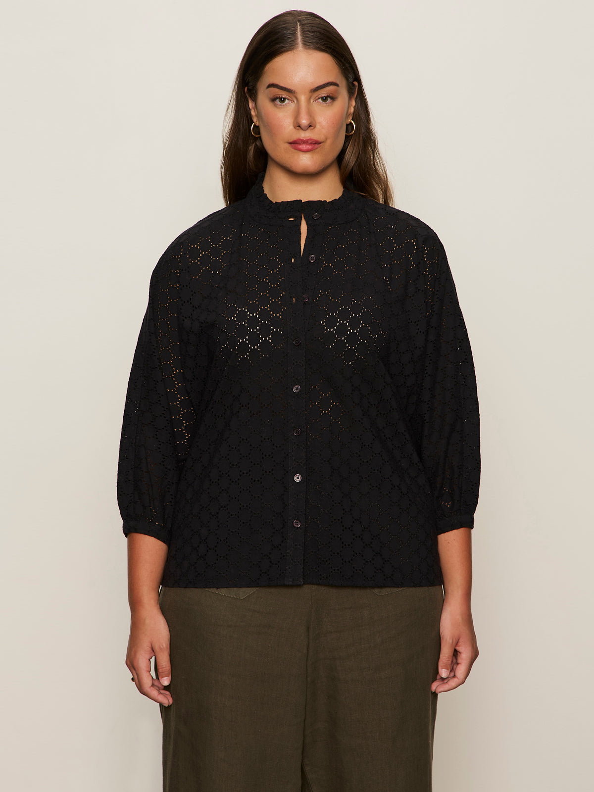A woman with long brown hair models the Sanctuary Clothing petal drop blouse in black (extended sizing), a loose-fitting, eyelet button-up with three-quarter sleeves, paired with olive green pants against a neutral background.