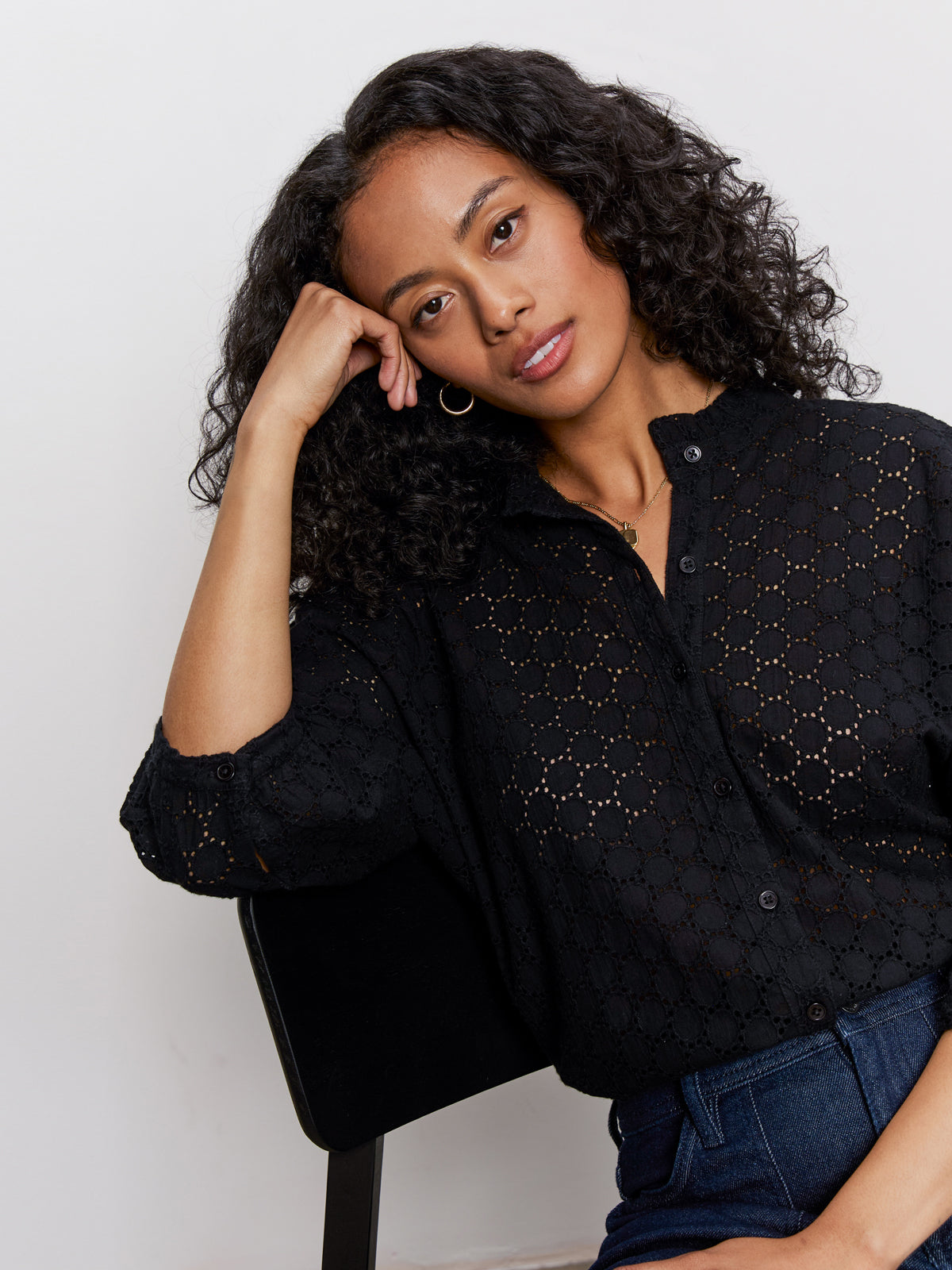 A woman with curly hair sits on a black chair, softly gazing at the camera. She wears blue jeans and the Sanctuary Clothing petal drop blouse in black.