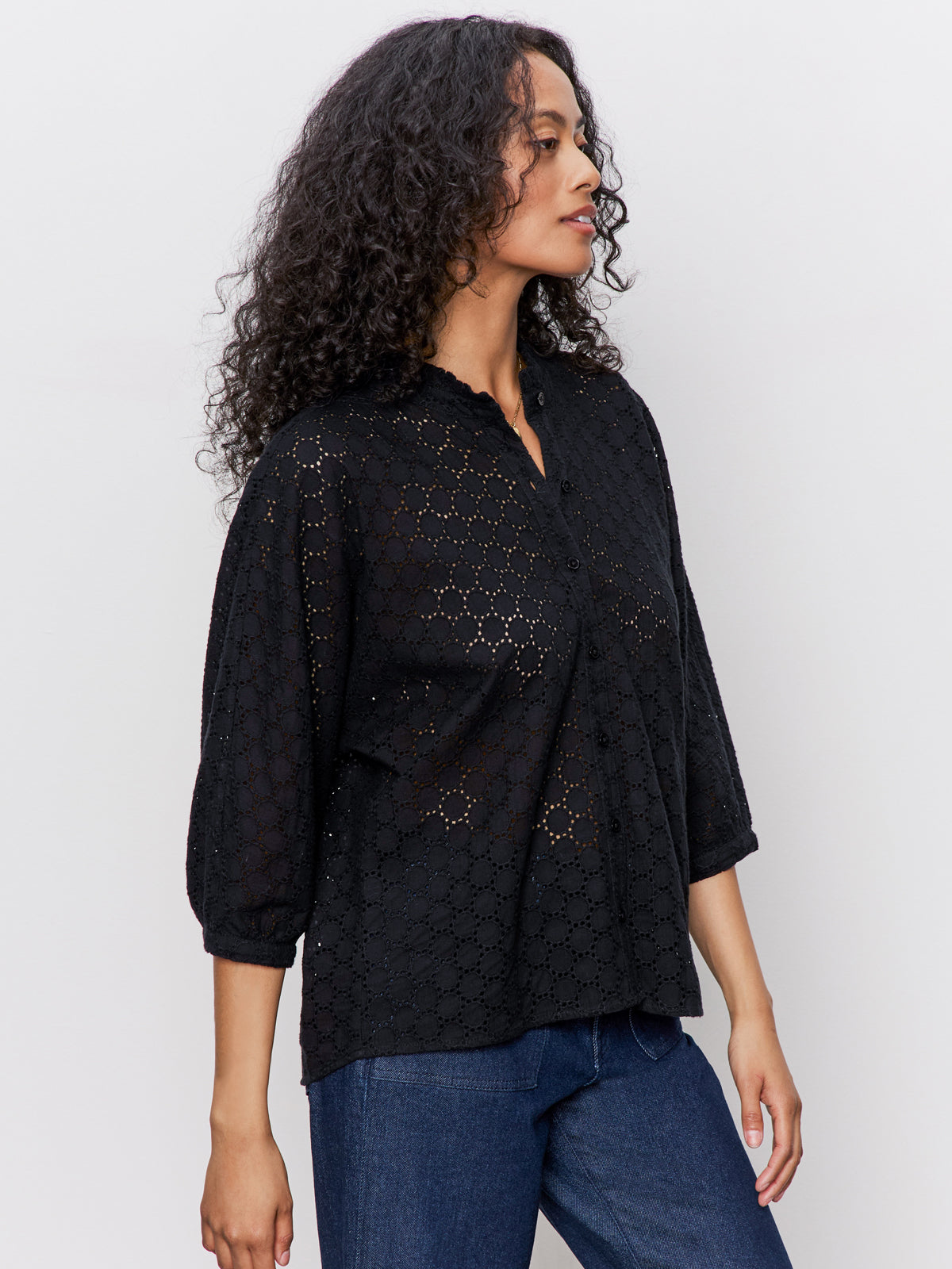A woman with long curly hair wears the Sanctuary Clothing petal drop blouse in black and blue jeans while standing sideways against a plain light background.